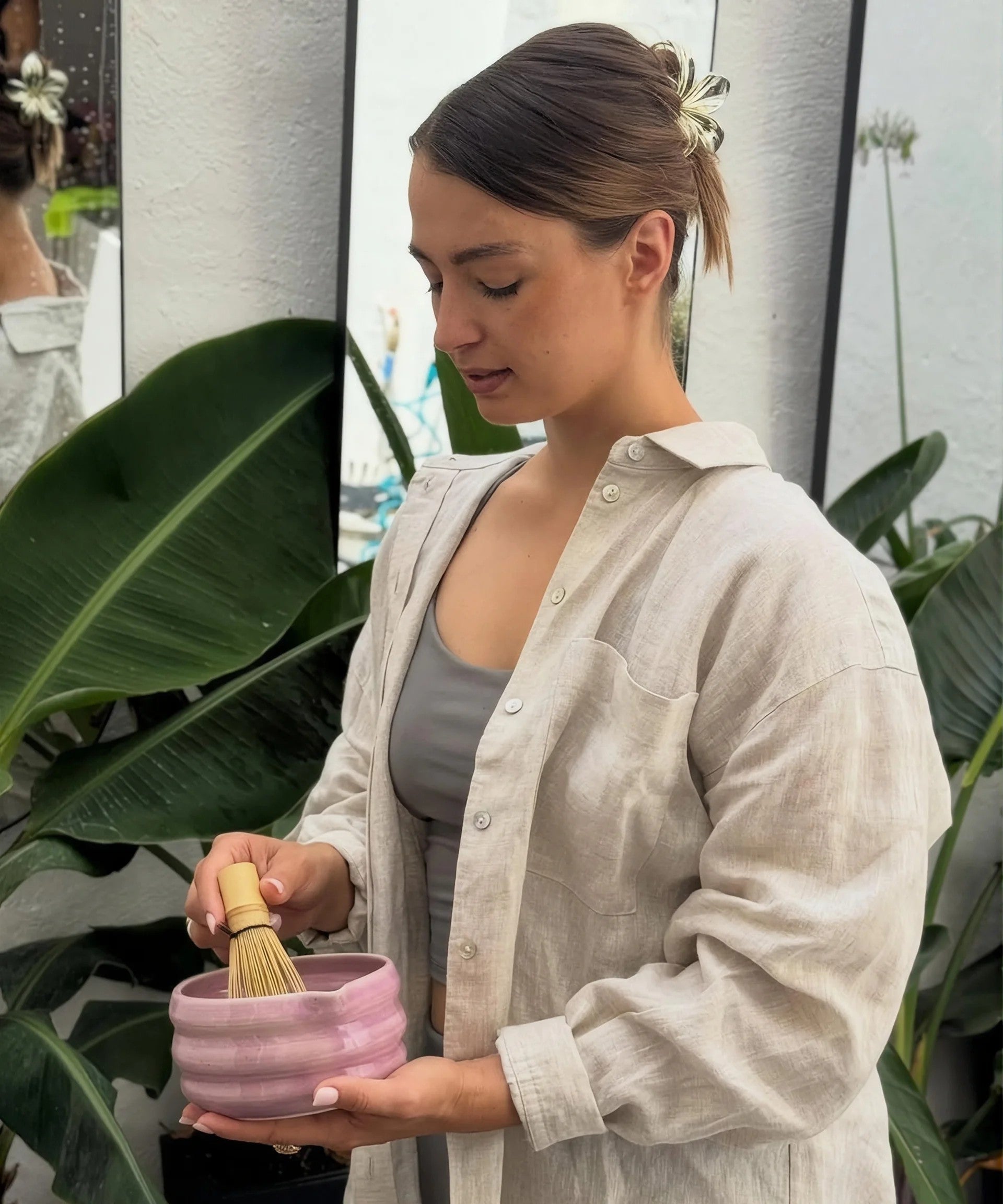 A woman in a linen shirt whisking matcha in a pink bowl among house plants